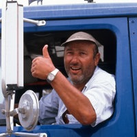 Manfred Krug in der Rolle des Truckers Franz Meersdonk während der Dreharbeiten zu "Auf Achse" in einem blauen Lastwagen. Er sitzt auf dem Fahrersitzt und lehnt sich aus dem Fenster, lächelt in die Kamera und streckt einen Daumen nach oben. 