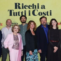 Ein Gruppenfoto des Casts des Films "Reich um jeden Preis" bei der Premiere auf dem roten Teppich. Die acht Schauspieler lächeln freundlich in die Kamera. Hinter ihnen an der gelben Wand steht der italienische Originaltitel "Ricchi a Tutti i costi".