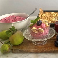 Im Hintergrund eine große Schale mit rosa Fruchtpüree und ein Blech mit Streuseln. Im Vordergrund eine kleine Schale mit Fruchtüree mit einer Pflaume verziert auf einem Holzbrett. Daneben ein Glas Pflaumensaft. Weitere Pflaumen und Birnen als Dekoration.