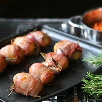 Kartoffelspieße mit Speck ummantelt auf einem schwarzen Blech. Danaben ein Rosmarinzweig. Im Hintergrund ein Topf gefüllt mit Paprika-Dip.