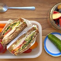 Von oben fotografiert: Eine Frischhaltedose mit einer aufgeklappten, belegten Stulle und Rohkost, daneben ein aufgeschraubtes Glas mit Obstsalat.