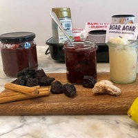 Drei Marmeladengläser mit roter und heller Marmeldade. Davor Zitronen, getrocknete Pflaumen und Zimtstangen, im Hintergrund weitere Zutaten in einer Box