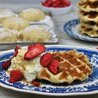 Eine Lütticher Waffel ist mit Sahne und Erdbeeren auf einem Teller angerichtet, im Hintergrund steht ein Teller mit weiteren Waffeln und ein Backblech mit Teig