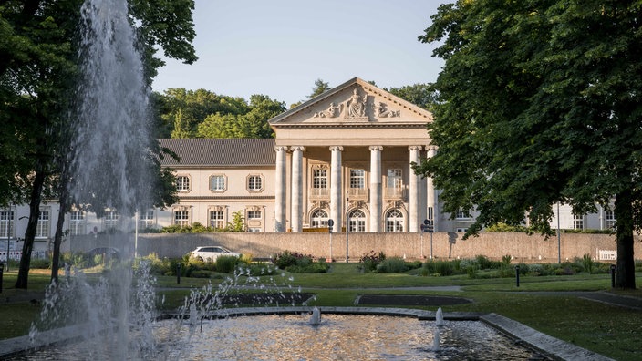 Das Bild zeigt das Aachener Kurhaus mit seinen Sandsteinwänden und dem überdachten Eingang. Das Dach wird von dicken Säulen getragen. Im Vordergrung sprenkelt ein Brunnen.