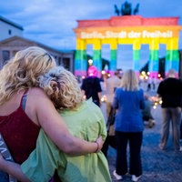 Zwei Menschen umarmen sich vor dem Brandenburger Tor, das in Regenbogenfarben angeleuchtet ist. 