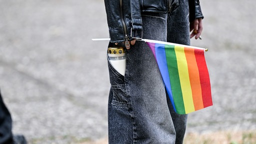 Eine Person mit einer Treuerkerze in der Hosentasche hält eine Pride-Flagge in der Hand