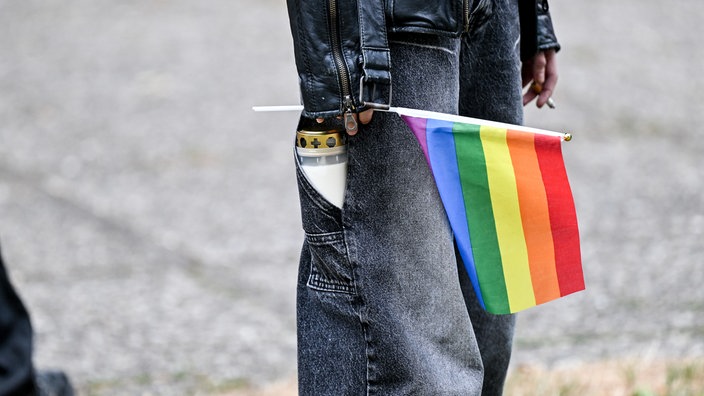 Eine Person mit einer Treuerkerze in der Hosentasche hält eine Pride-Flagge in der Hand