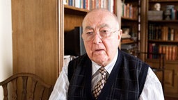Doktor Dressler hält ein Exposé in der Hand. Er überlegt, wie er seine Ersparnisse in Sicherheit bringen kann.