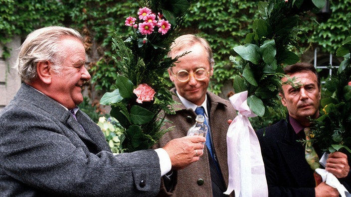 Egon (Wolfgang Grönebaum, links) will sich das "Spalier-Stehen" mit Schnaps versüßen. Hans (Joachim Hermann Luger, Mitte) und Enrico (Guido Gagliardi) sind wenig begeistert von dieser Idee.
