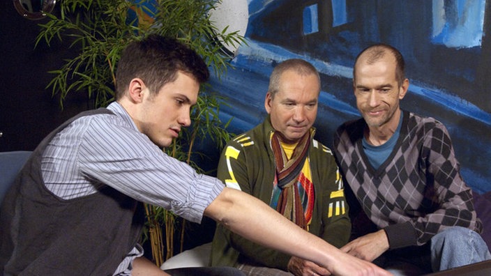Felix (Oliver Moser, links) besucht seine Väter „Käthe“ (Claus Vinçon, Mitte) und Carsten (Georg Uecker). Er zeigt ihnen Fotos seiner Reisen durch Kanada.