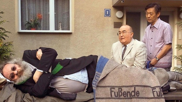 Für Harry (Harry Rowohlt) hält Dressler (Ludwig Haas, rechts daneben: Amorn Surangkanjanajai) einen guten Rat bereit. Nur für seinen eigenen Sohn Frank hat der pensionierte Arzt nichts übrig.