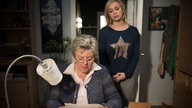 Helga liest an Leas Schreibtisch, während diese hinter ihr steht