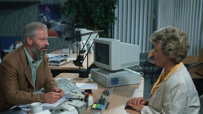 Helga (Marie-Luise Marjan) lässt sich im neuen Reisebüro von Erich Schiller (Bill Mockridge) Vorschläge für ihren Urlaub machen.