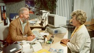 Helga sitzt bei Erich Schiller im Reisebüro und lässt sich Vorschläge für ihren Urlaub machen