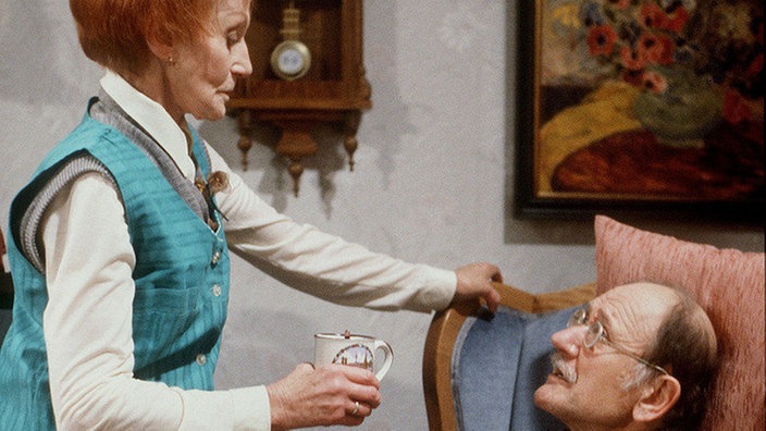 Hubert (Robert Zimerling) nutzt seine Erkältung weidlich aus und lässt sich von Rosi (Margret von Munster) bedienen.