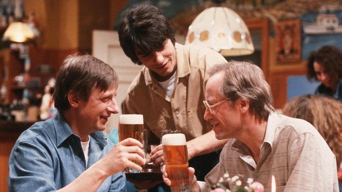Im "Akropolis" treffen Franz (Raimund Gensel, links) und Hans (Joachim Hermann Luger) aufeinander. Vasily (Hermes Hodolides, Mitte) serviert ihnen gerne ein Bier.