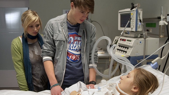 Lea (Anna-Sophia Claus, links) und Nico (Jeremy Mockridge) stehen traurig am Krankenbett: Caro (Cynthia Cosima) liegt noch immer im Koma.