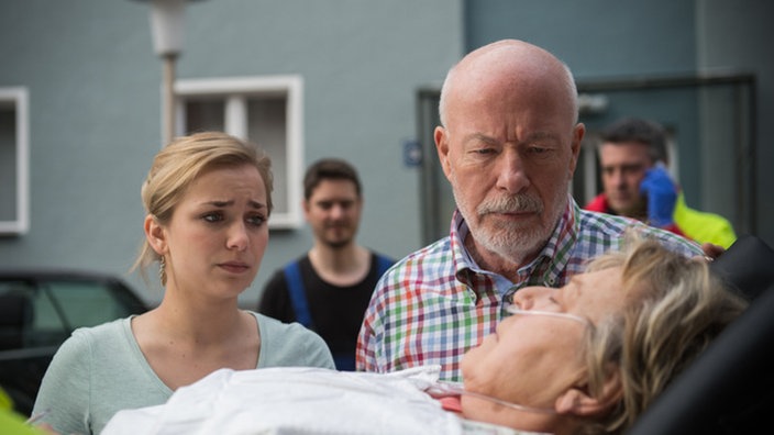 Lea und Erich schauen besorgt zu Helga auf einer Trage