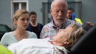 Lea und Erich schauen besorgt zu Helga auf einer Trage