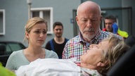 Lea und Erich schauen besorgt zu Helga auf einer Trage