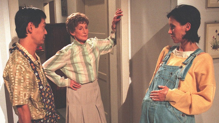 Rosi (Margret van Munster, Mitte), Urszula (Anna Nowak) und Gung (Amorn Surangkanjanajai) beraten, wie sie die Wohnung für Huberts Rückkehr schmücken sollen.