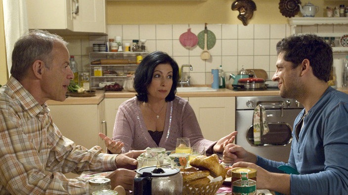 Sabrina (Susanna Capurso) will Enzo (Toni Snetberger, rechts) überreden, sich noch einmal krank schreiben zu lassen – dann könnte er länger bei ihr und Scholz (Knut Hinz) in München bleiben.