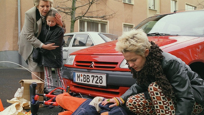 Tom liegt auf der Straße und Pat, Helga und Sarah stehen daneben