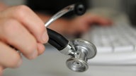 Ein Arzt hält mit der einen Hand ein Stethoskop, mit der anderen tippt er auf einer Computertastatur