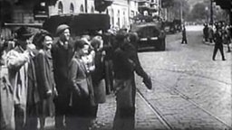 Ein schwarz-weiß-Foto von einer alten Strasse, im Vordergrund stehen menschen, im Hintergrund sieht man Panzer fahren