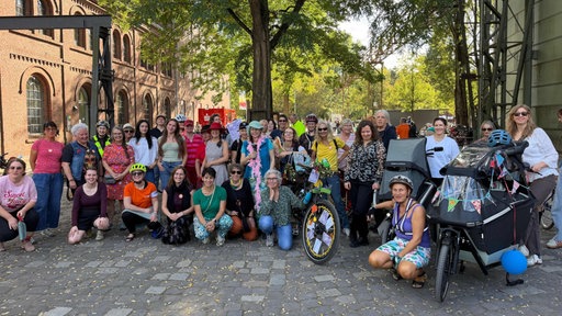 Eine Gruppe von Frauen, einige mit Fahrradhelm oder Fahrrad, stehen oder hocken draußen.