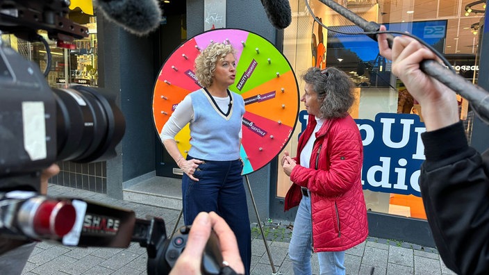 Moderatorin Lisa Ortgies im Gespräch mit einer Frau vorm Glücksrad beim PopUp-Studio