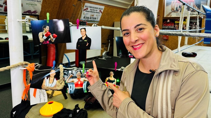 Die Ex-Boxweltmeisterin Dilar Kisikyol steht in einer Sporthalle neben einem Boxring und zeigt lächelnd auf Fotos, die sie in ihrer Boxkarriere zeigen.