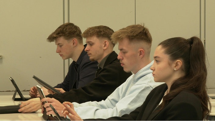 Vier Polizeischüler:innen sitzen in einem Klassenraum nebeneinander, vorne eine junge Frau, dahinter drei junge Männer und arbeiten an Tablets.