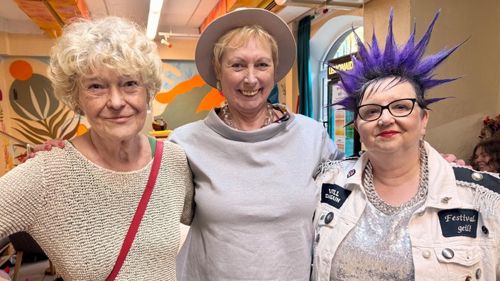Drei Frauen stehen eng beieinander und lächeln in die Kamera. Eine trägt einen Hut, eine hat lockige graue Haare, die dritte auffällige violette Irokesenfrisur und eine silberne Jacke.