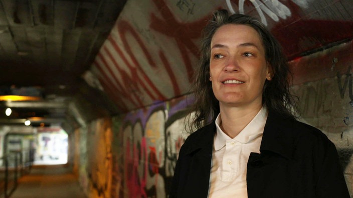 Die Architektin Karin Hartmann steht in einer mit Graffiti bemalten Unterführung in Bonn. Sie trägt eine weiße Bluse und eine dunkle Jacke und blickt lächelnd zur Seite.
