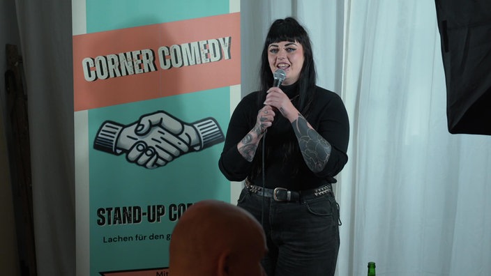 Xenia mit Mikrofon auf der Bühne, im Hintergrund ist ein Schild, auf dem "Corner Comedy" steht