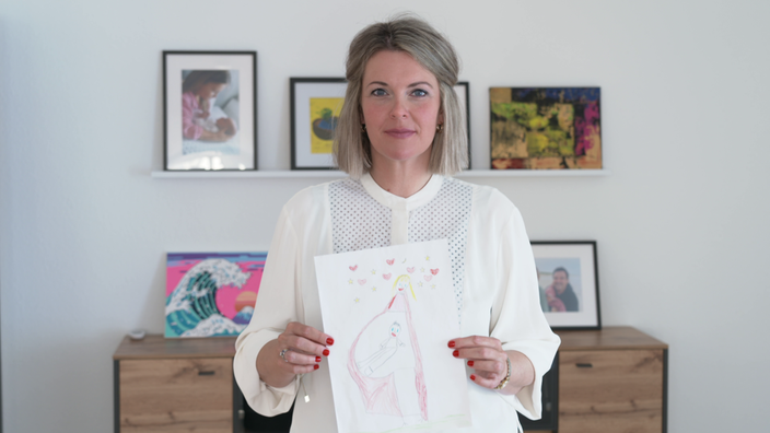 Porträt einer Frau mit blonden Haaren, weißer Bluse und gemaltem Kinderbild, das sie in die Kamera hält.