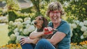 Katrin Iskam sitzt mit einem Hund auf dem Arm auf einer Wiese und strahlt in die Kamera.