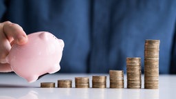 Eine Person hält ein Sparschwein über aufgetürmte Euromünzen. Symbolbild: Geldanlage, Finanzbildung