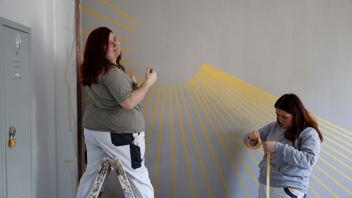 Eine Jugendliche (links) steht in Arbeitskleidung vor einer grauen Wand mit gelben Klebestreifen. Eine andere Jugendliche kniet rechts.