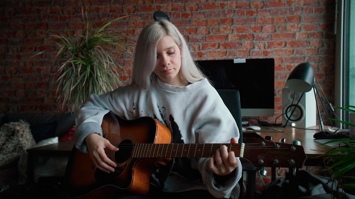 Eine blonde Frau mit grauen Pulli spielt Gitarre in einem Zimmer mit Backsteinwand.