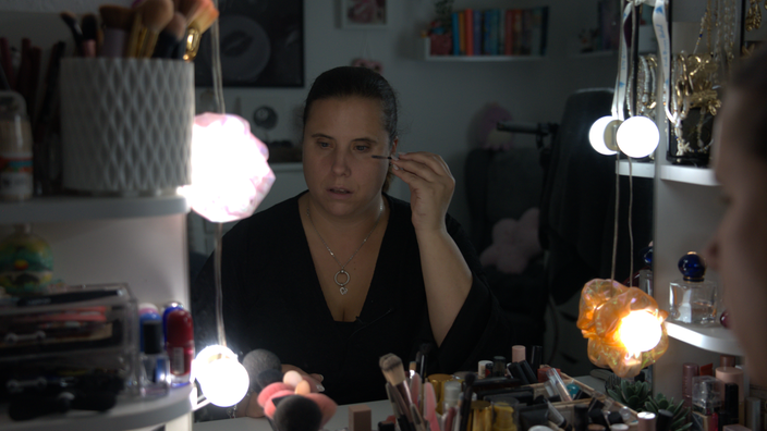 Frau schminkt sich im Spiegel. Sie trägt Wimperntusche auf. Um sie herum sind Schminkutensilien.