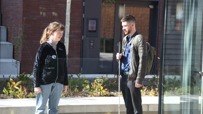 Rechts steht ein blinder Mann mit Stock. Links eine Frau, die zu dem Mann schaut.