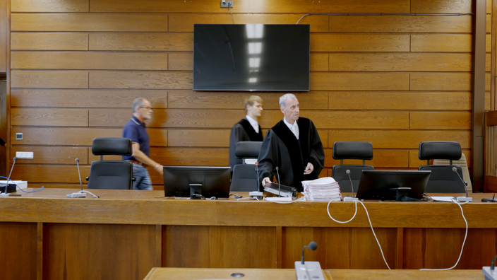 Das Richterpult in einem Gerichtssaal bestehend aus einem langem Tisch mit zwei Bildschirmen, sowie vier Stühlen und zwei Männern in Richterkutte und einem in Jeans und Shirt.