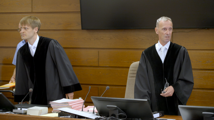 Zwei Männer in Richterkutte stehen vor dem Richterpult in einem Gerichtssaal.