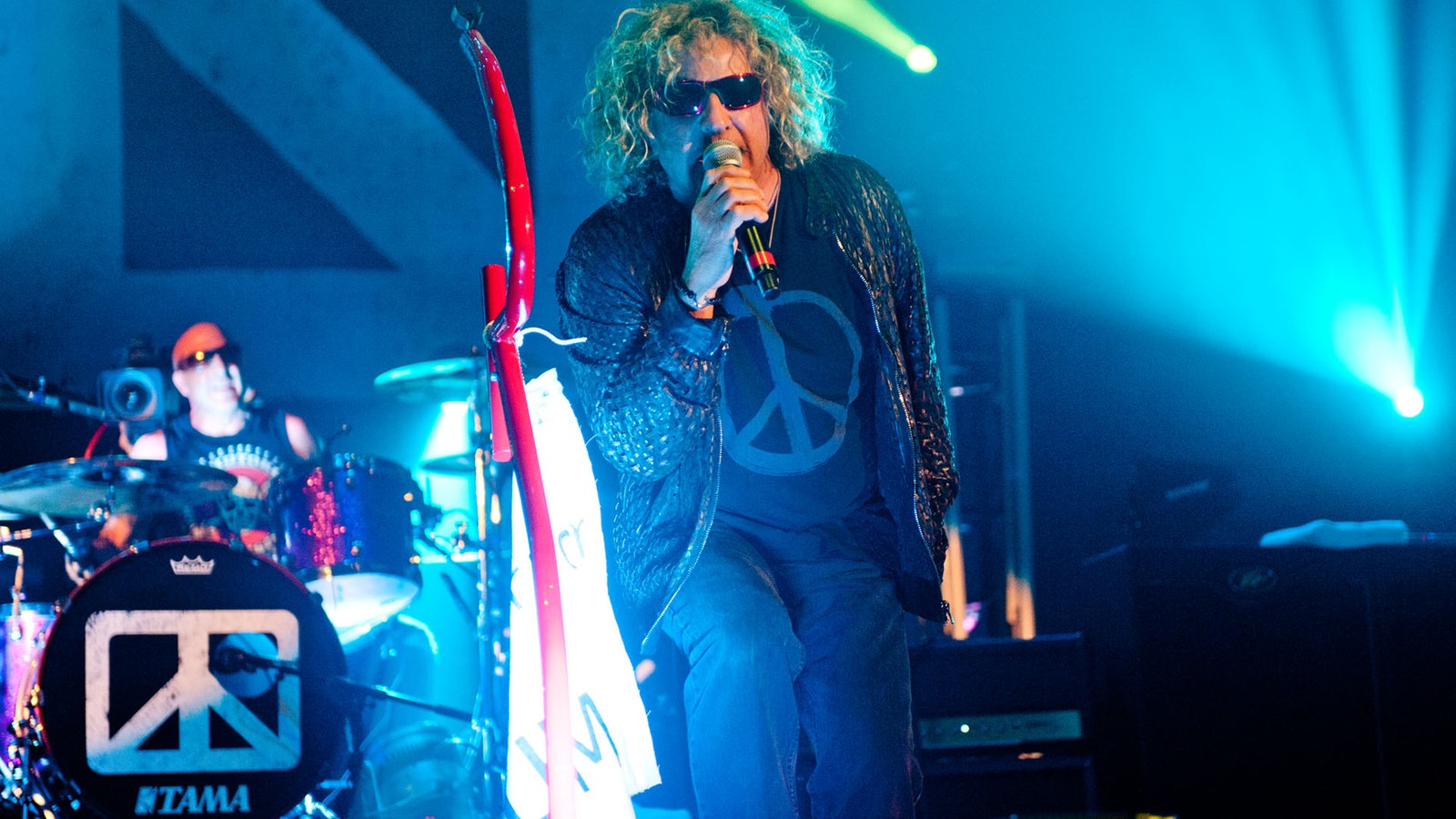 Sammy Hagar, der Sänger der Chickenfoots hält sich mit der rechten Hand das Mikrofon an den Mund. Mit dem rechten Bein stützt er sich ab und lehnt sich zum Publikum nach vorne