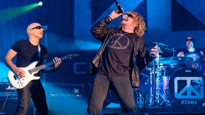 Sammy Hagar lehnt sich mit dem Mikrofon am Mund zurück links steht Joe Satriani an seiner Gitarre und im Hintergrund ist der Drummer an seinem Schlagzeug zu sehen