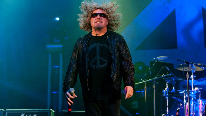 Sammy Hagar springt wodurch seine Haare hoch fliegen. In der rechten Hand hält er sein Mikrofon