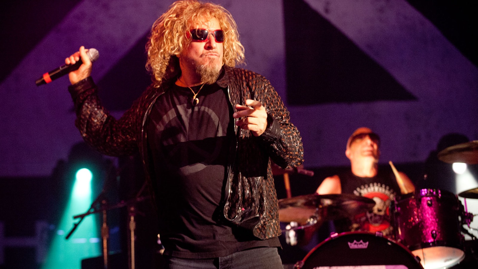 Sammy Hagar hat den rechten Arm mit dem Mikrofon in der Hand nach oben angewinkelt mit der linken Hand zeigt er in Richtung Publikum