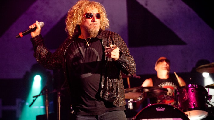 Sammy Hagar hat den rechten Arm mit dem Mikrofon in der Hand nach oben angewinkelt mit der linken Hand zeigt er in Richtung Publikum